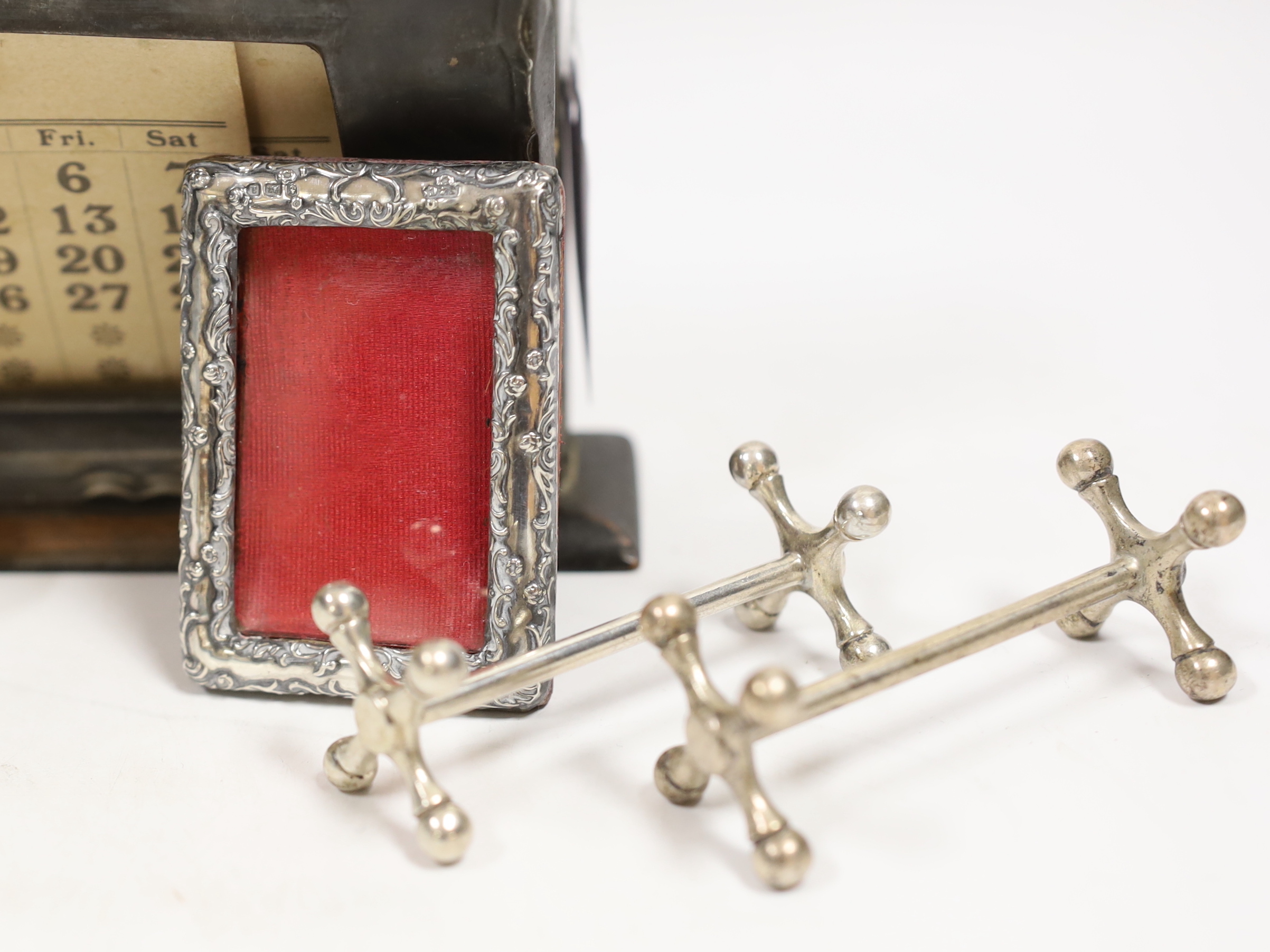 A pair of Edwardian silver knife rests, William Hutton & Sons, London, 1908, 77mm, a silver mounted wooden desk calendar, a late Victorian small silver mounted photograph frame, a silver mounted glass posy vase and a whi